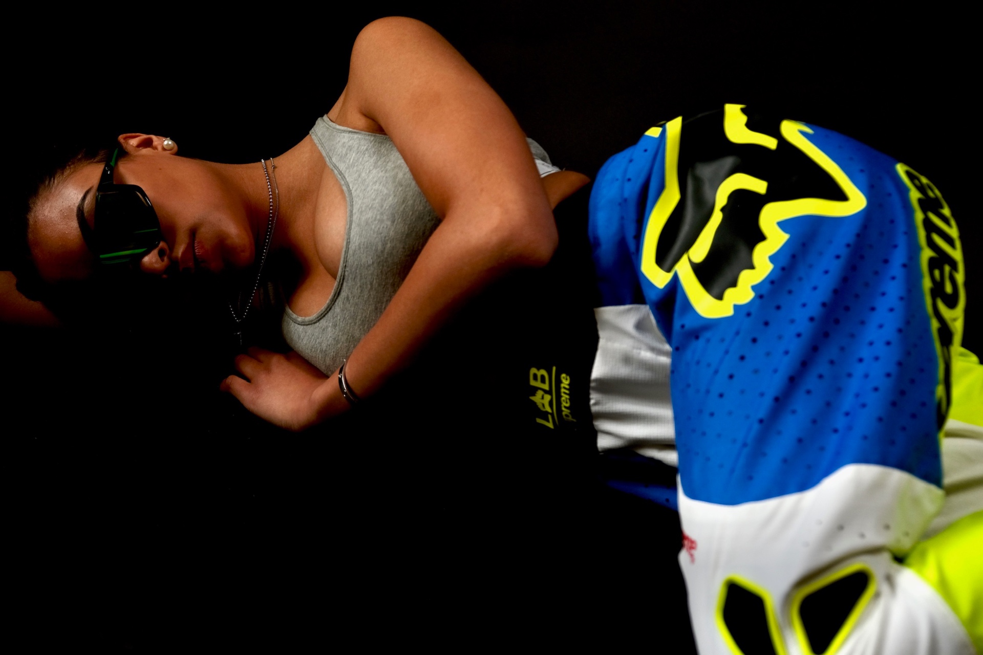 Woman in sunglasses lying beside a motocross jersey against a black background