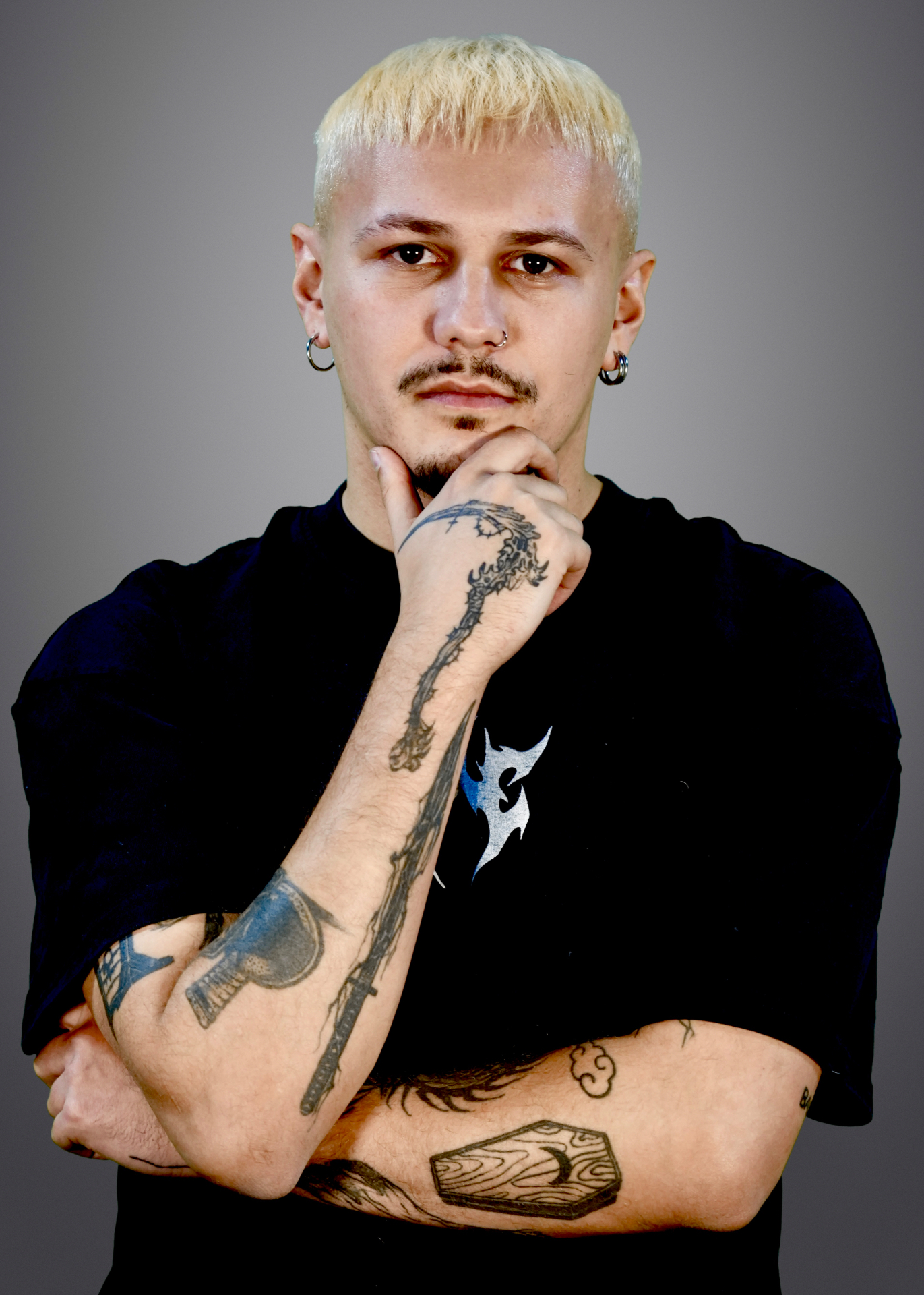 Studio portrait of a person with bleached hair, tattoos, and a black shirt, hand resting on chin