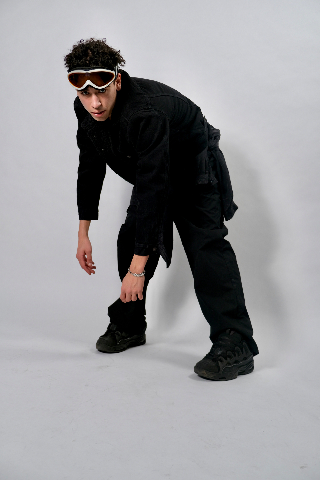 Studio portrait of a man in all-black clothing and ski goggles leaning forward